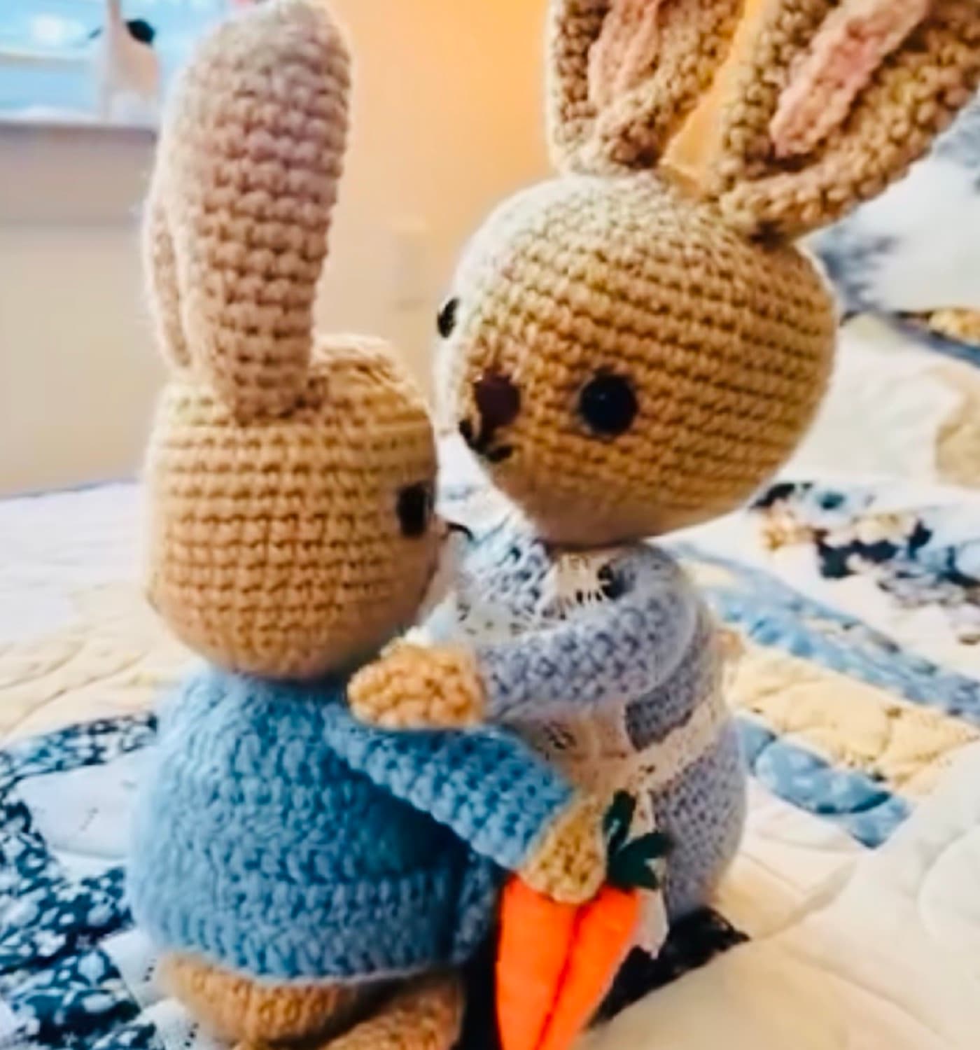 Two crocheted bunnies in blue jackets sharing a felt carrot on a quilted bedspread